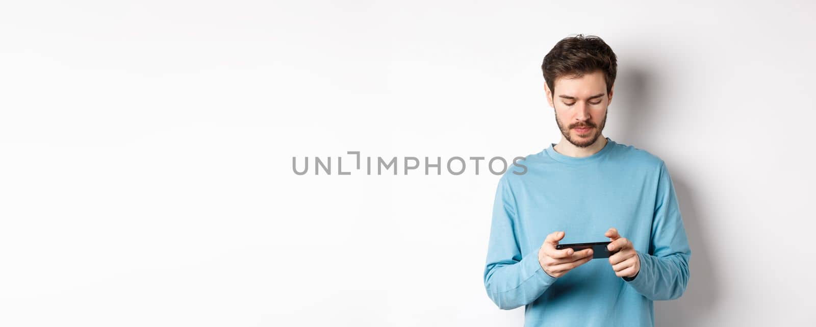 Male model playing video game on smartphone, looking serious at mobile screen, standing in sweatshirt over white background.