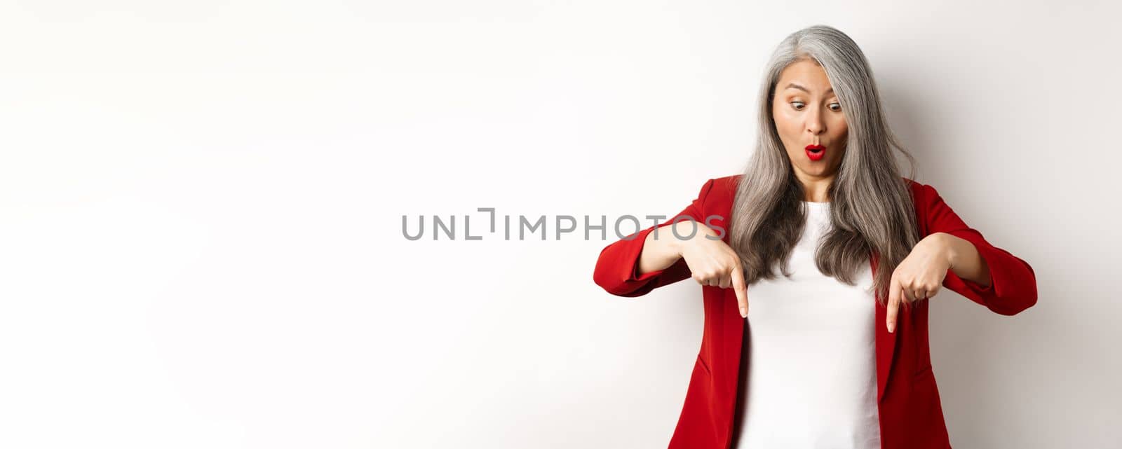 Impressed asian mature woman checking out promo, pointing and looking down with amazed face, standing in blazer over white background.