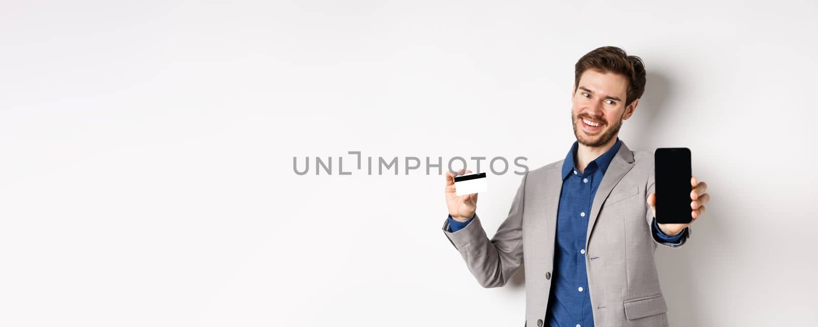 Online shopping. Successful businessman holding plastic credit card and showing mobile screen, smiling satisfied, making money, white background.
