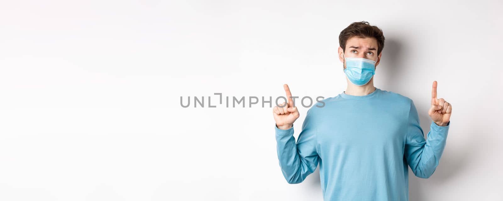 Coronavirus, health and quarantine concept. Skeptical and doubtful man in medical mask pointing, looking up, feeling indecisive, standing over white background by Benzoix