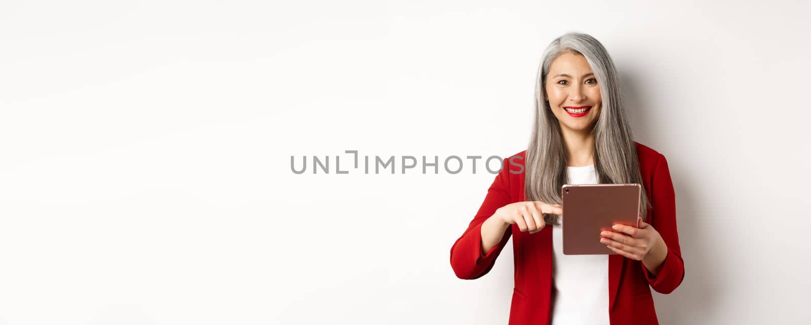 Business. Smiling senior businesswoman using digital tablet, pointing finger at screen, standing over white background.