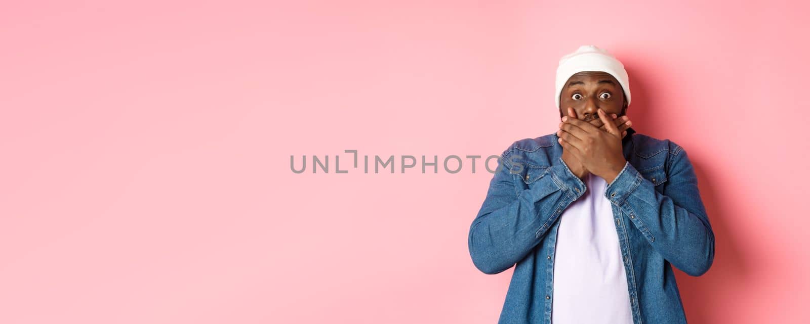 Shocked african-american man cover mouth and staring at camera, standing speechless against pink background.