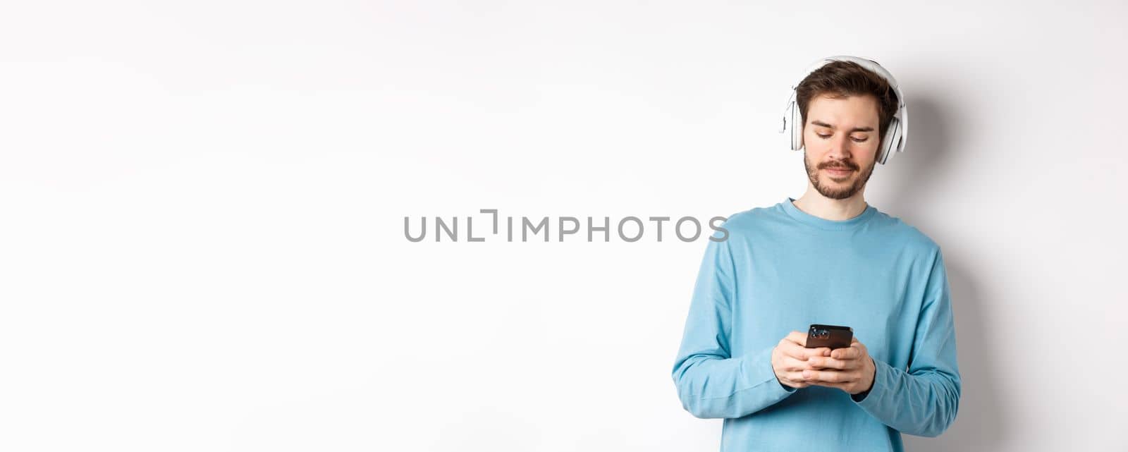 Modern young man in blue sweatshirt put on music on smartphone, listening songs in wireless headphones, white background.