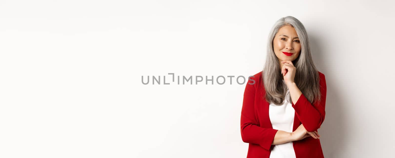 Business concept. Asian mature businesswoman smiling pleased, looking thoughtful, having an idea, standing in red blazer over white background.