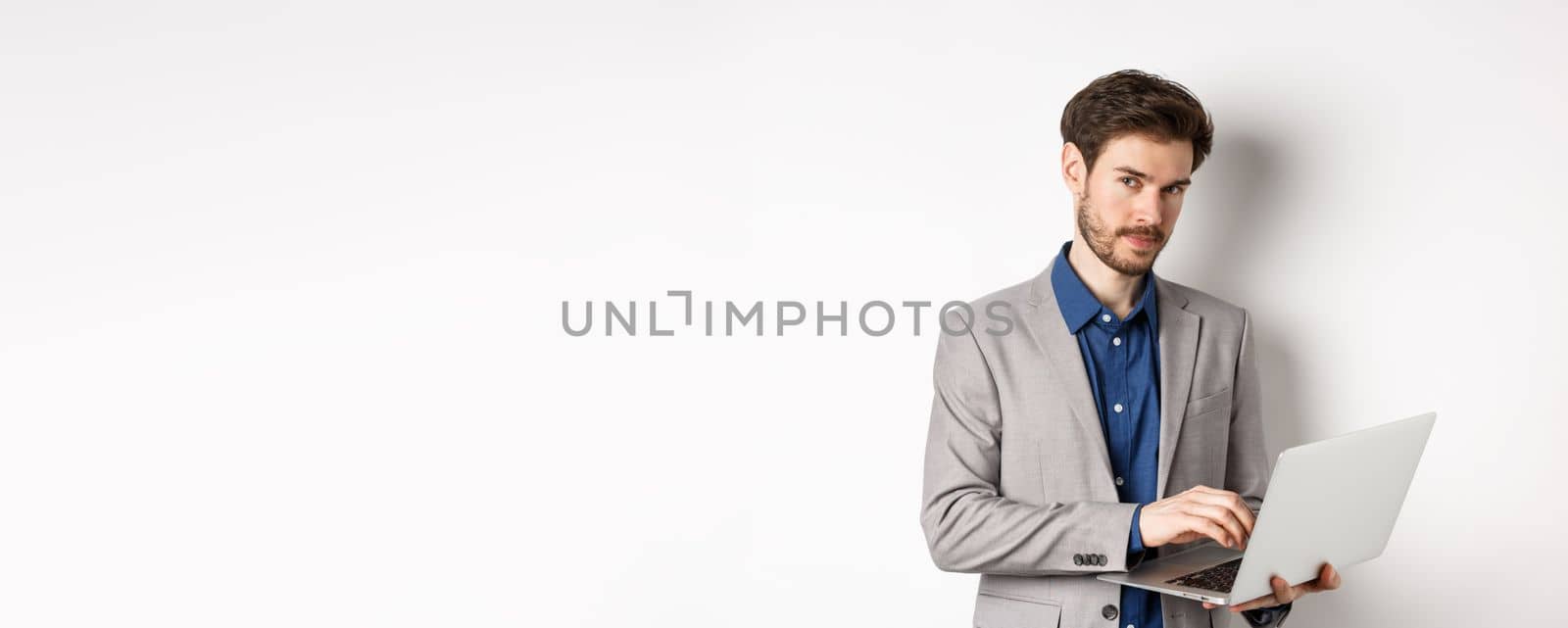 Handsome businessman in suit working on laptop, looking at camera confident, standing against white background.