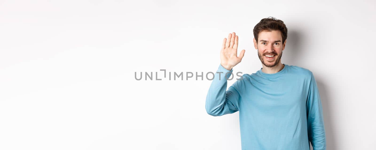 Cheerful young man with beard saying hello, looking friendly and waving hand to greet you, standing over white background.