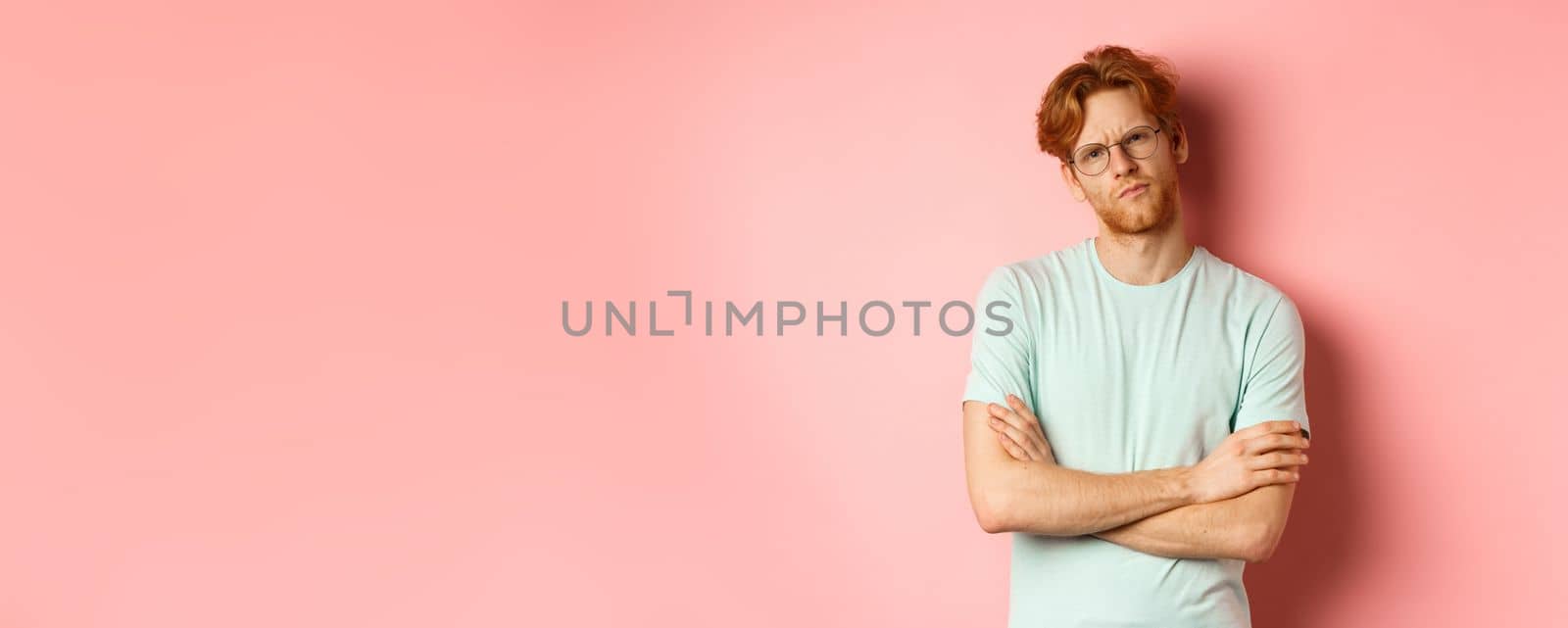 Young man with red hair and beard, wearing glasses and t-shirt, cross arms on chest, frowning while staring with skeptical and doubtful expression, standing over pink background.