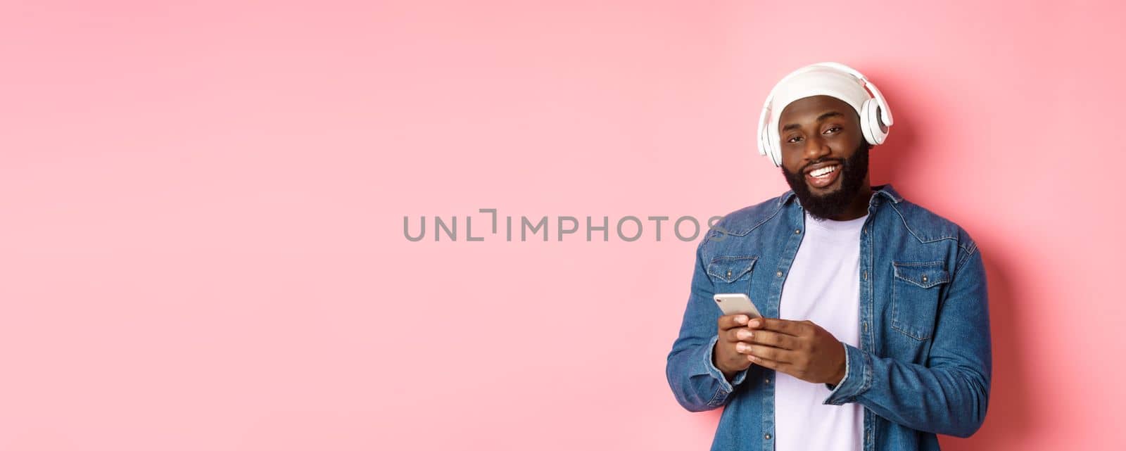 Handsome hipster guy in headphones smiling satisfied at camera, listening music in headphones, using mobile app, standing over pink background.