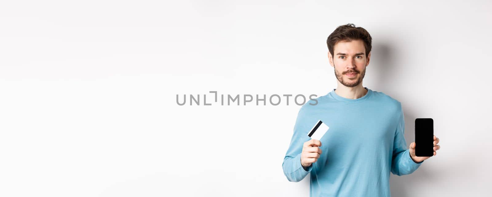 Handsome caucasian man showing empty smartphone screen and plastic credit card, standing over white background.