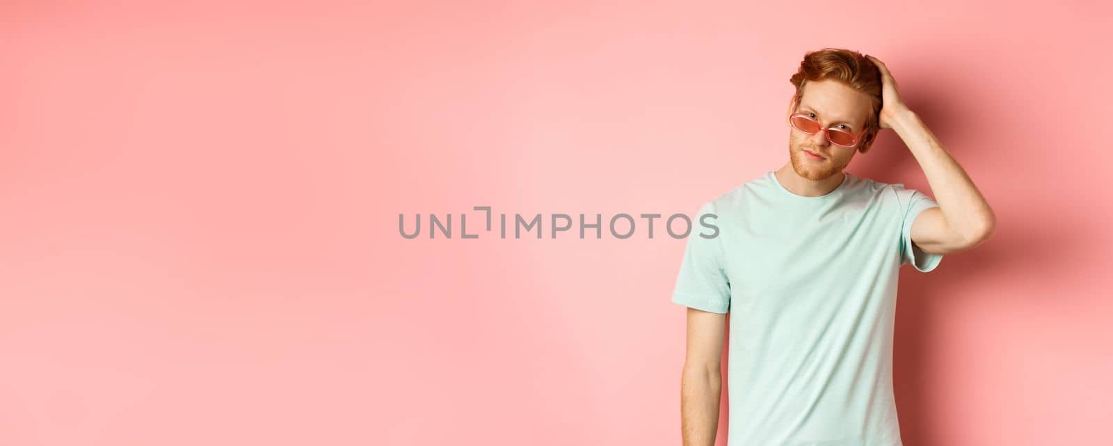 Handsome young redhead man in sunglasses, brushing hair with hand and looking smug and confident at camera, standing over pink background.