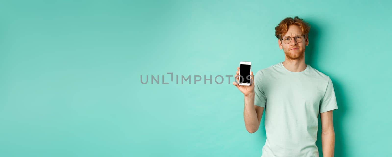 Skeptical male model with red hair and glasses, showing mobile screen and frowning disappointed, dislike application, standing over turquoise background.