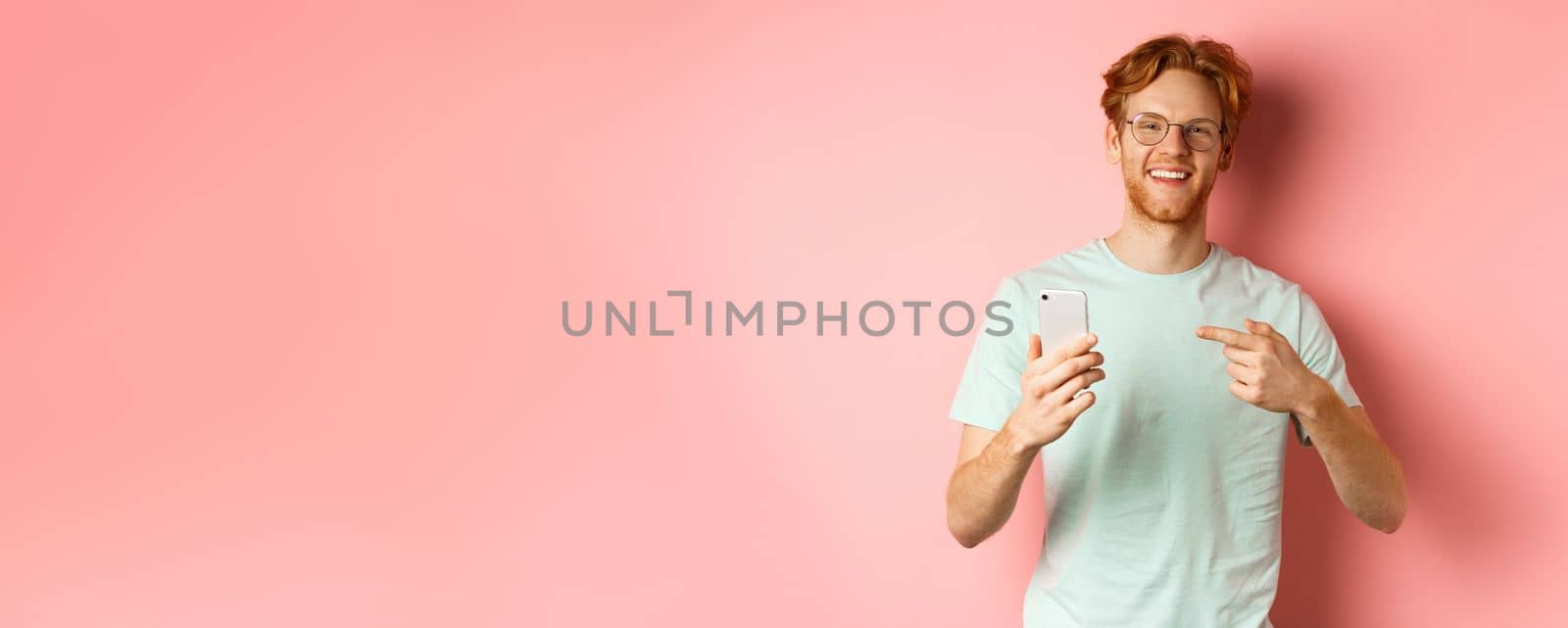 Young man with red hair and beard, wearing t-shirt and glasses, smiling while pointing finger at smartphone, recommend online promotion, pink background.