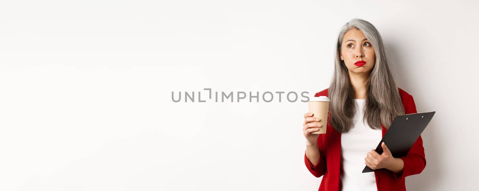 Tired asian female office worker holding clipboard and paper cup, drinking coffee and exhaling with exhausted face, standing over white background.