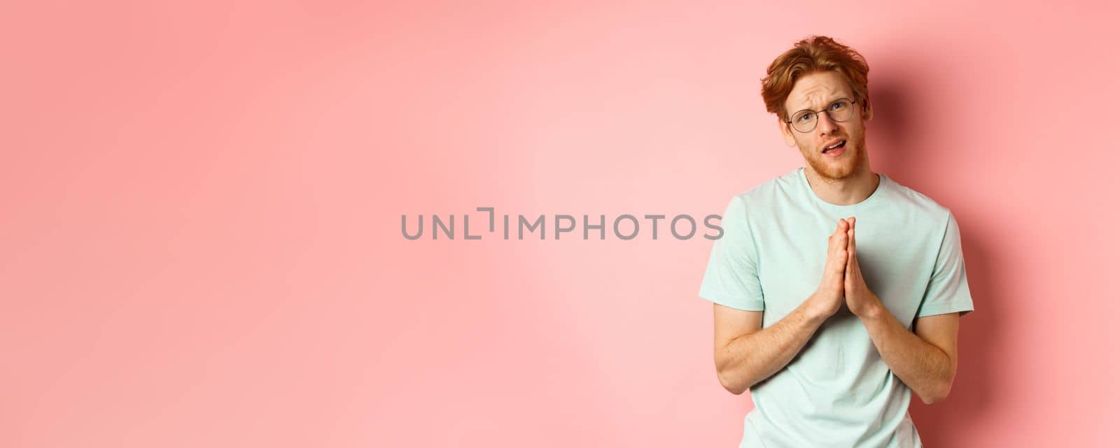 Redhead bearded guy begging for favour, holding hands in namaste gesture and asking for help, need something, standing over pink background.