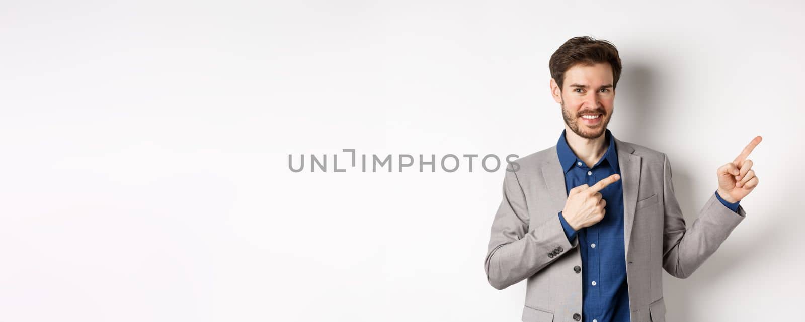 Handsome smiling man in business suit pointing fingers at upper right corner, showing logo, advertising company, standing on white background.