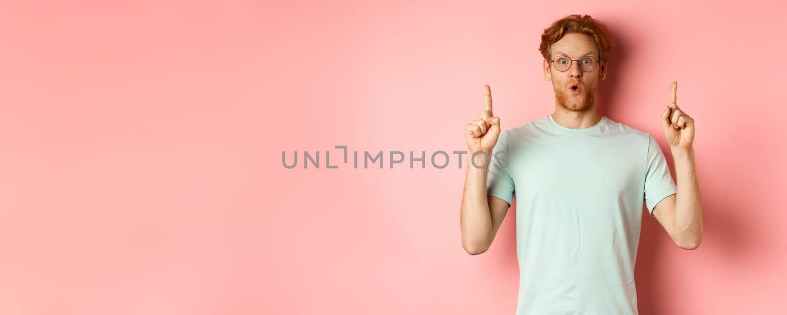 Impressed caucasian man with ginger hair, wearing glasses and t-shirt, saying wow and pointing fingers up at awesome deal, standing over pink background by Benzoix