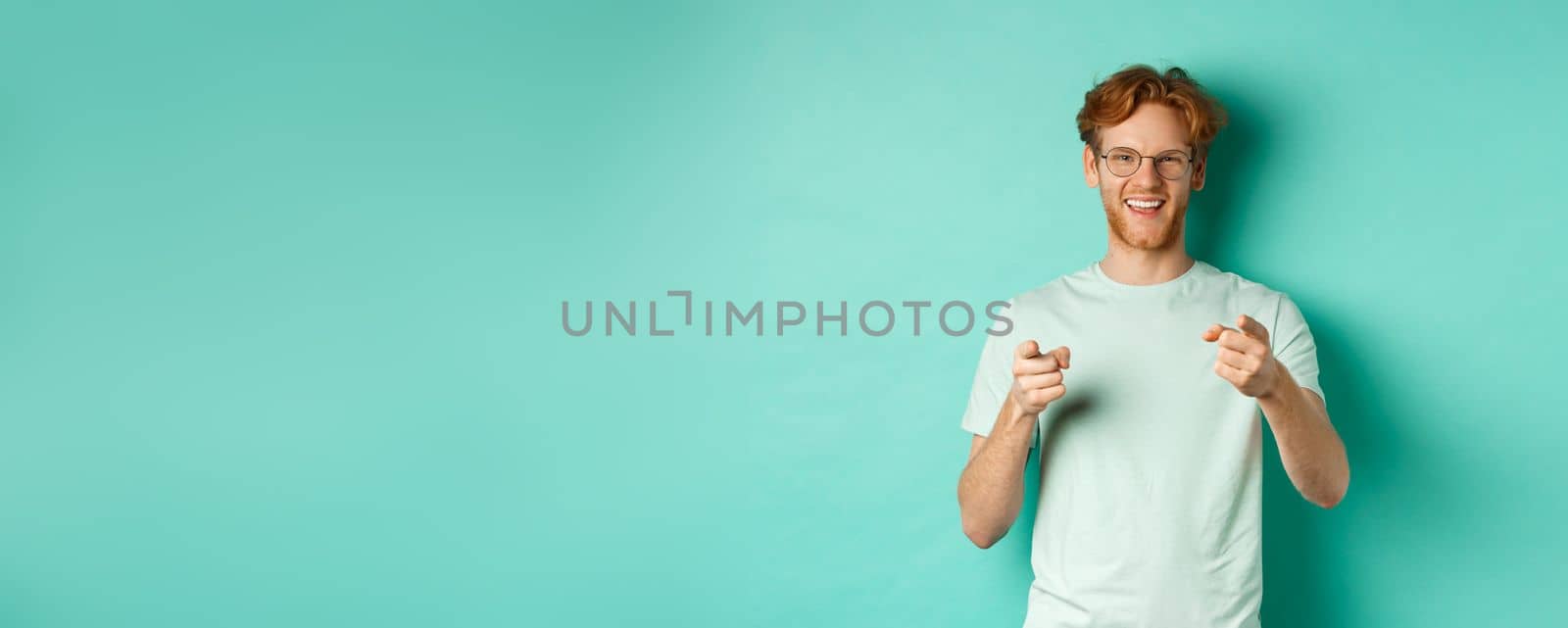 Handsome young man with ginger hair, wearing glasses and t-shirt, pointing finger at camera and smiling, choosing you, congratulating or praising, standing over mint background.