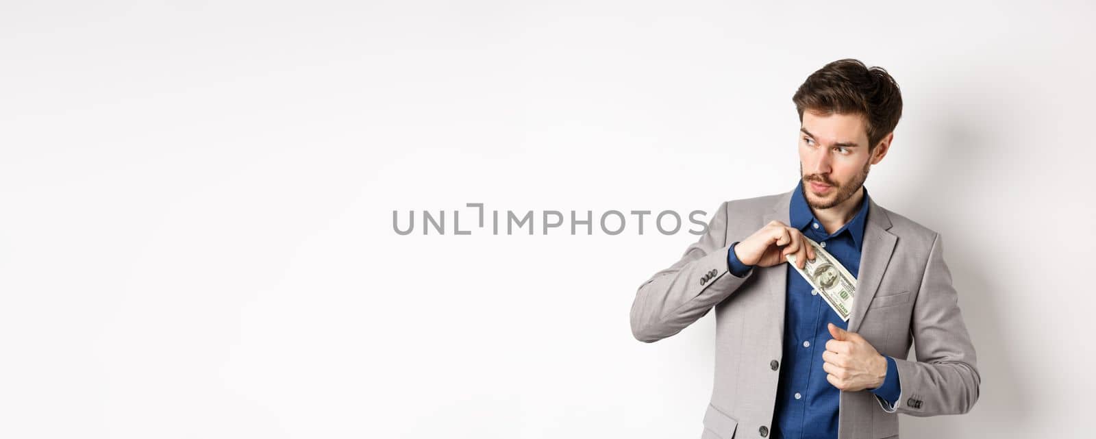 Confident businessman put money in suit and looking aside, standing on white background.