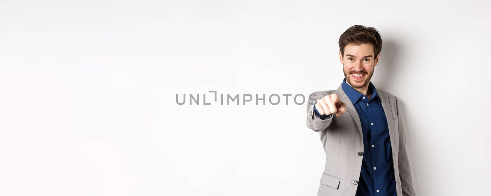 Its you. Smiling handsome manager in suit pointing at camera, choosing person and looking pleased, standing on white background.