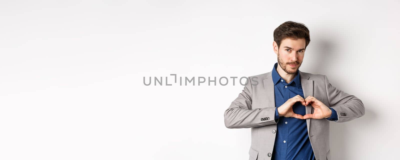 Romantic man in suit showing heart sign and smiling, love his girlfriend, standing on white background.