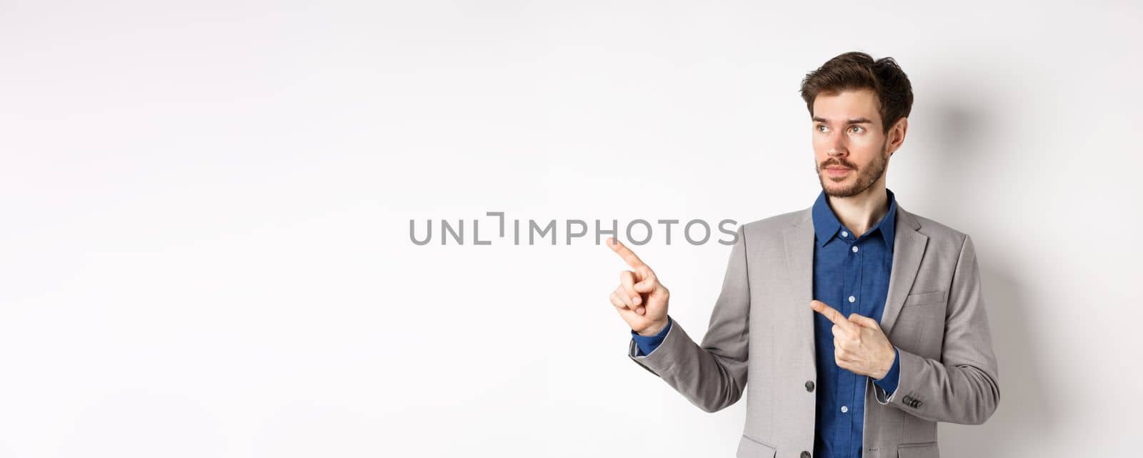 Handsome businessman in suit pointing, looking left at logo with serious face, standing against white background.