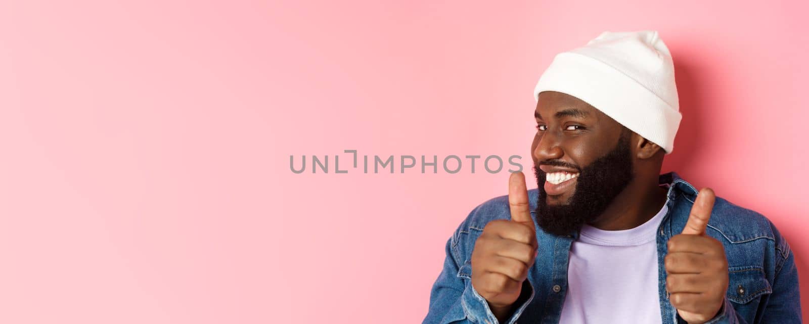 Close-up of happy Black bearded guy in beanie showing support, agree or approve something, giggle devious and showing thumbs-up, standing over pink background.