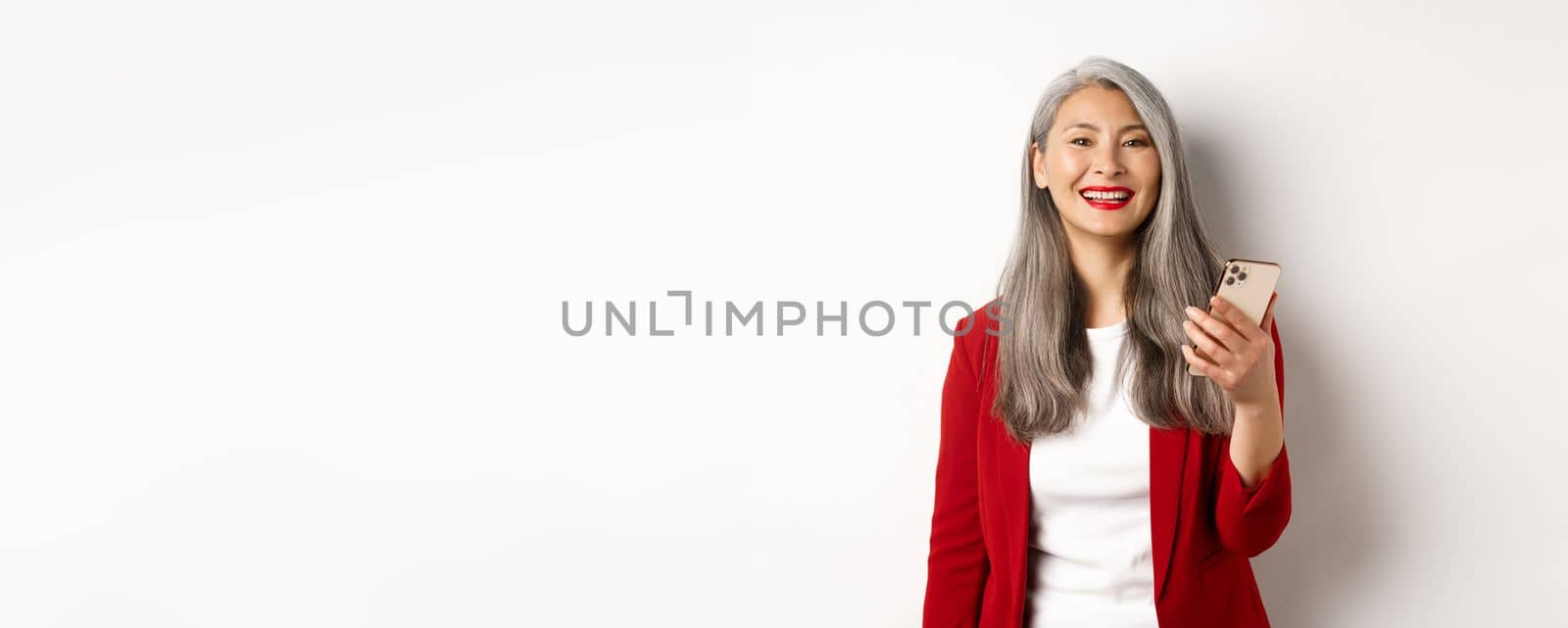 Asian female entrepreneur in red blazer using smartphone, smiling happy at camera, standing over white background.
