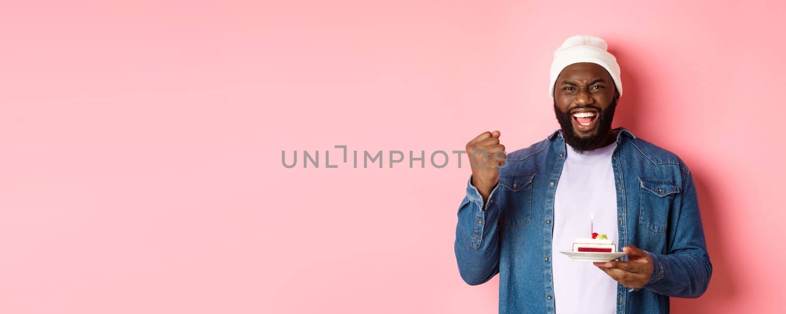 Cheerful african-american guy celebrating birthday, making wish on bday cake with lit candle, smiling happy, standing over pink background.