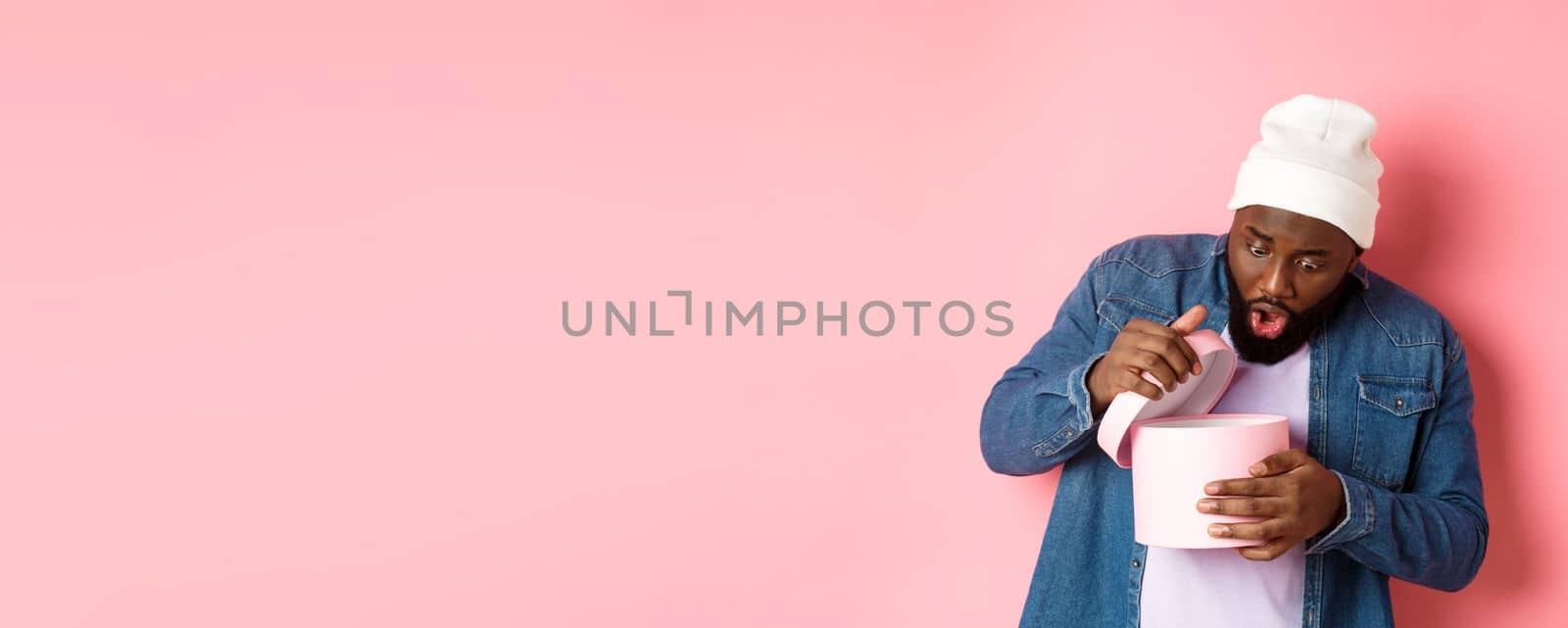 Image of surprised african-american man open box with birthday gift, staring at present with amazement, standing against pink background by Benzoix