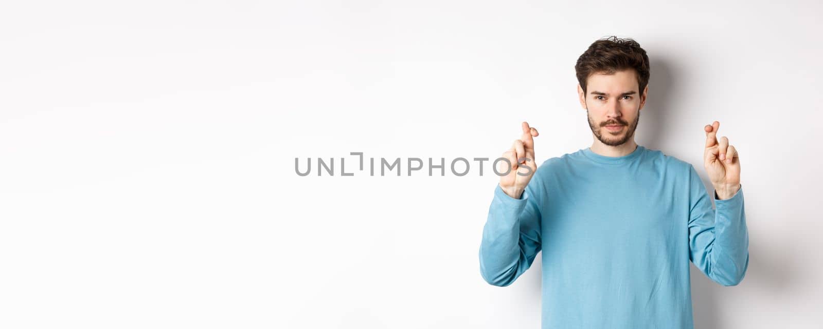 Hopeful and confident young man cross fingers for good luck, making wish or praying, anticipating results, standing over white background.
