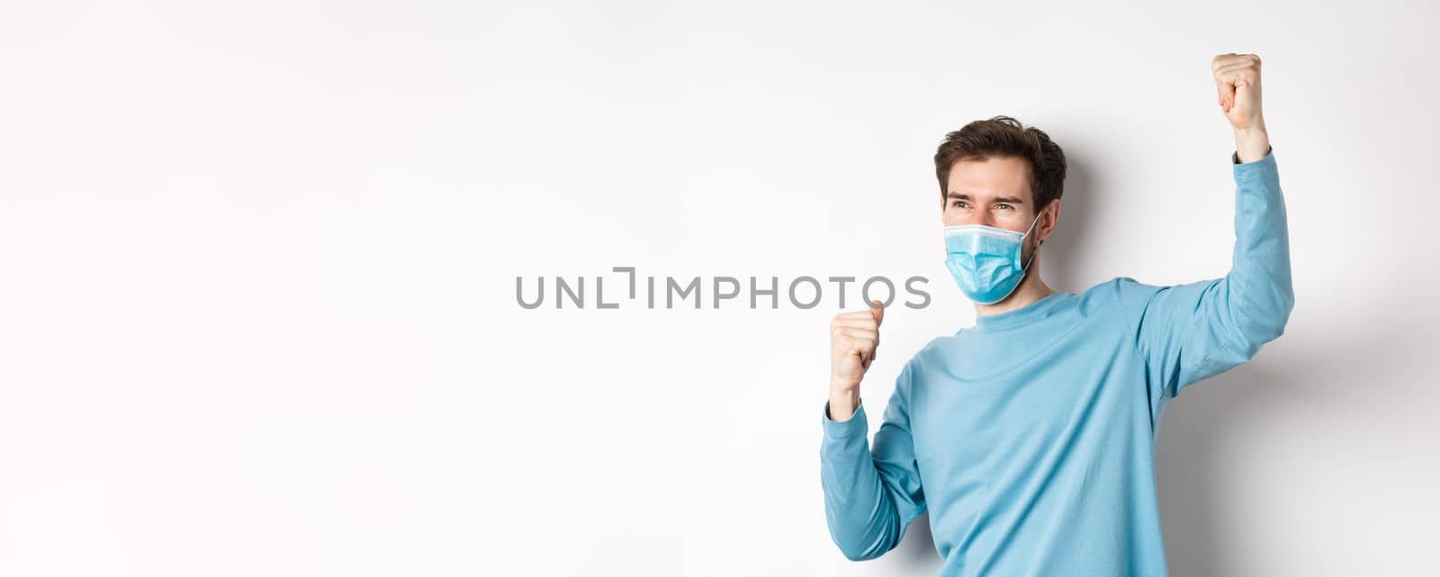 Covid-19, pandemic and social distancing concept. Happy man in medical mask watching sports game and cheering, raising hands up to celebrate, standing over white background.