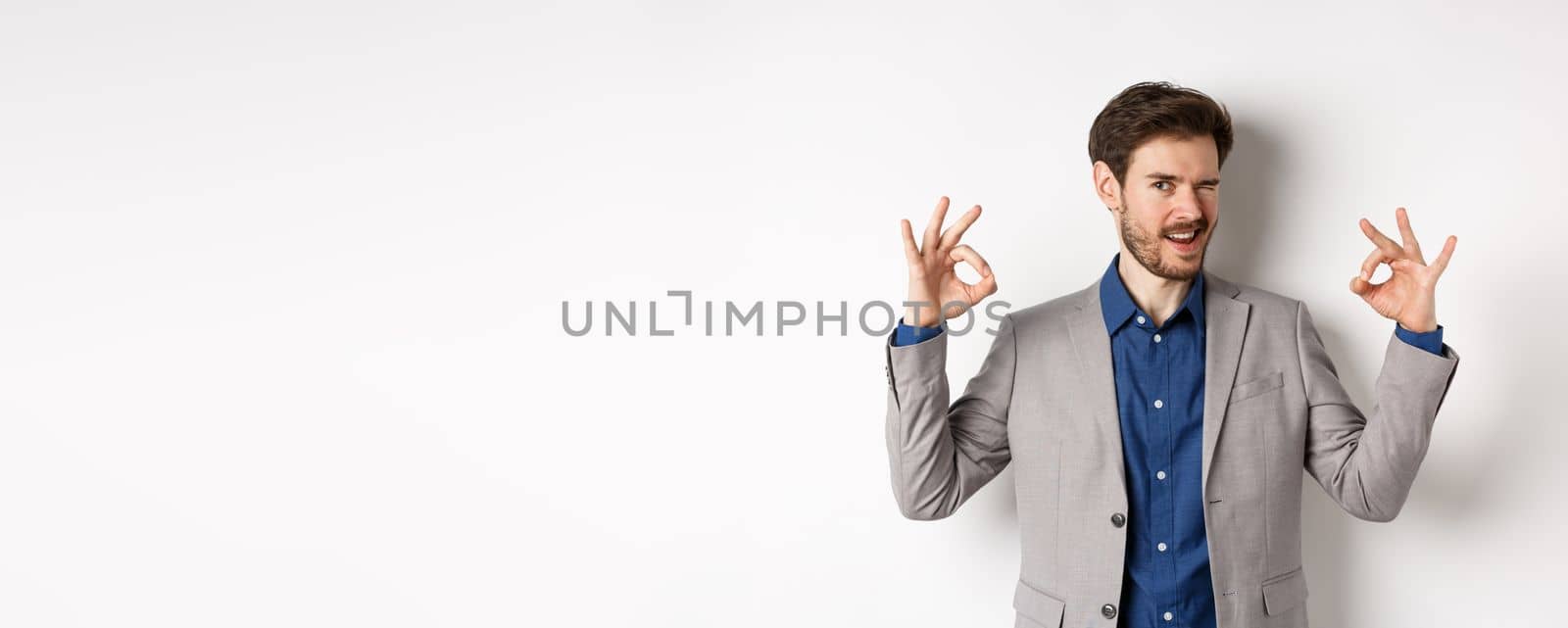 Cheeky handsome man in suit winking at you, showing okay signs, all under control gesture, approve good choice, assuring everything OK, standing on white background.