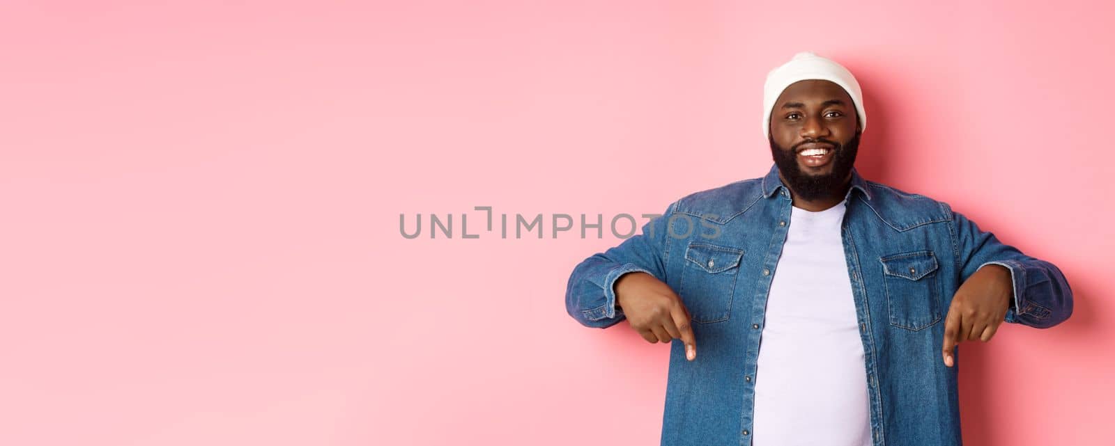 Handsome hipster african-american man pointing fingers down, smiling and showing promo offer, standing over pink background.