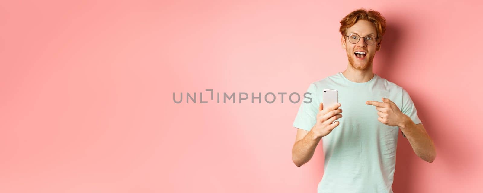 Cheerful guy talking about internet promo, smiling amazed and pointing finger at smartphone, standing over pink background by Benzoix