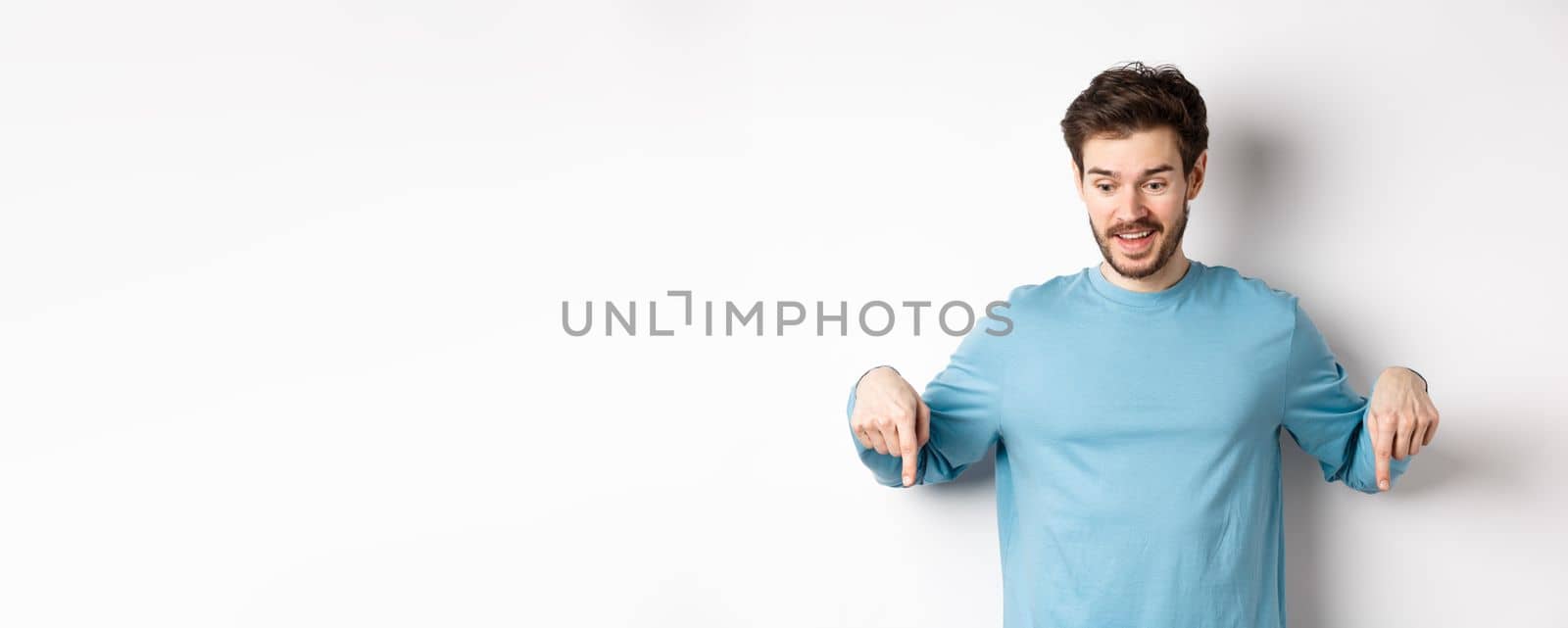 Excited and happy young man looking and pointing down at awesome deal, checking out promotion offer, standing on white background.