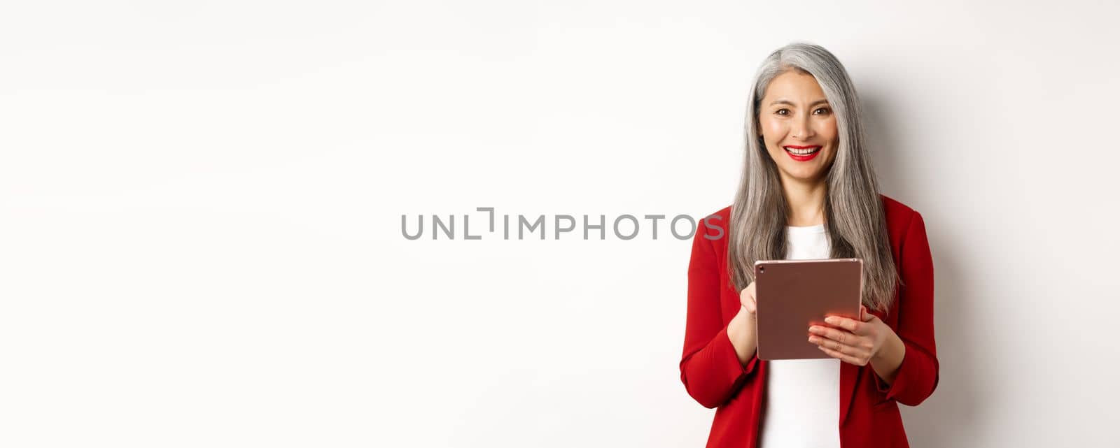 Business. Senior female entrepreneur working on digital tablet and smiling happy at camera, wearing red blazer and makeup, standing over white background by Benzoix