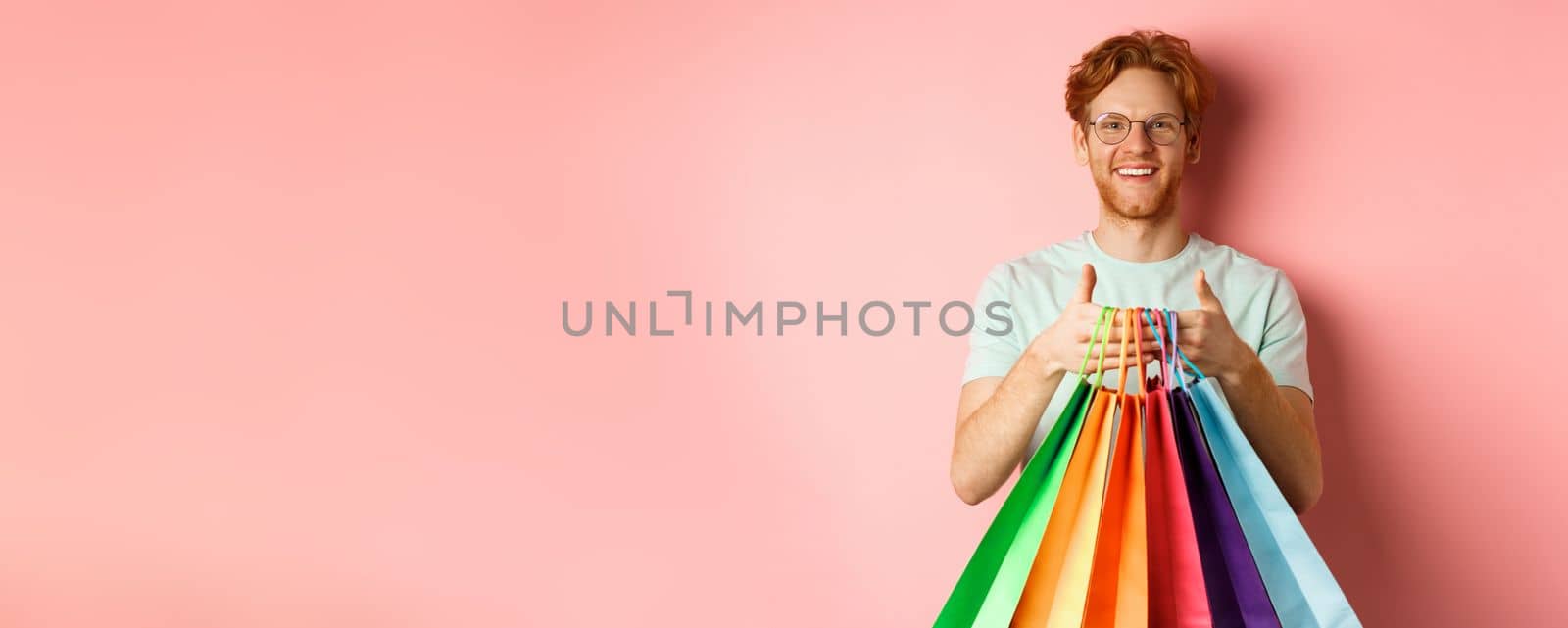 Cheerful redhead man buying gifts, holding shopping bags and smiling, standing over pink background by Benzoix