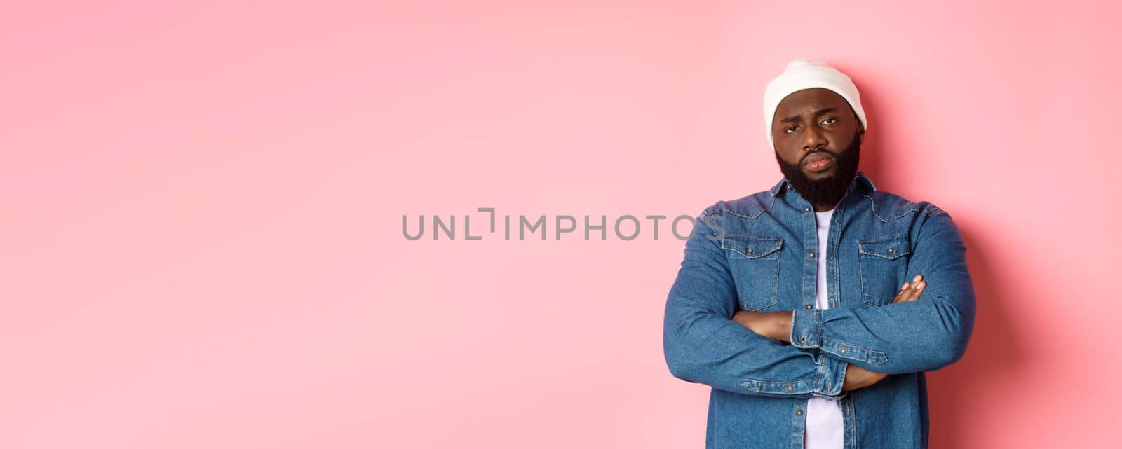 Disappointed african-american man in hipster beanie, staring at camera displeased, cross arms on chest, standing over pink background by Benzoix