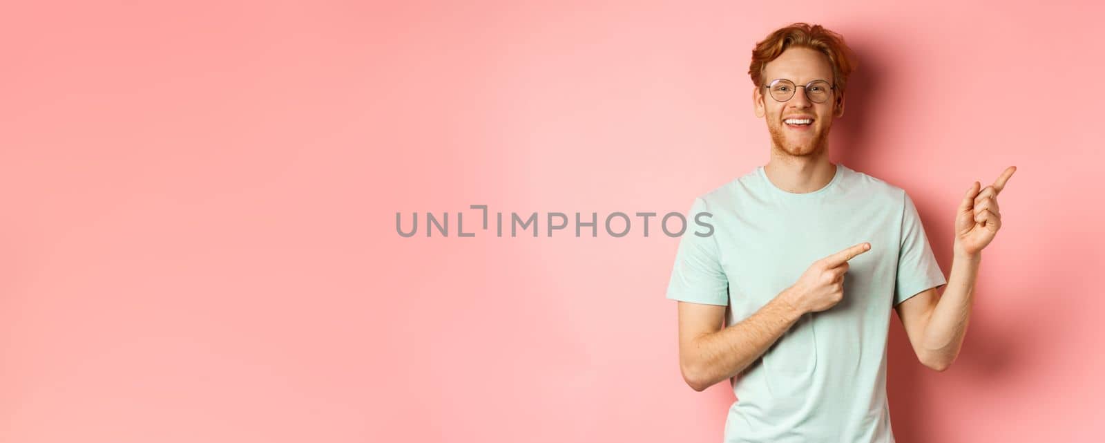 Friendly redhead guy in glasses pointing at upper right corner, showing promo banner and smiling at camera, standing over pink background by Benzoix