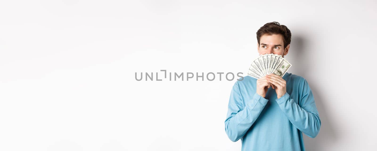 Dreamy man thinking of buying product, holding money and looking left pensive, imaging shopping, standing over white background.