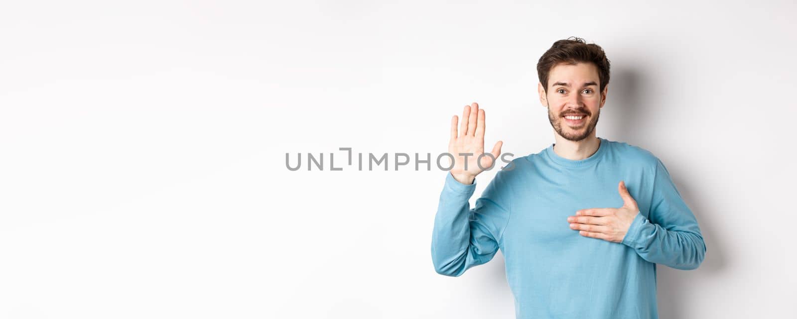 Smiling hosent man raising arm and hold hand on heart, making promise tell truth, swear or give oath, standing over white background.