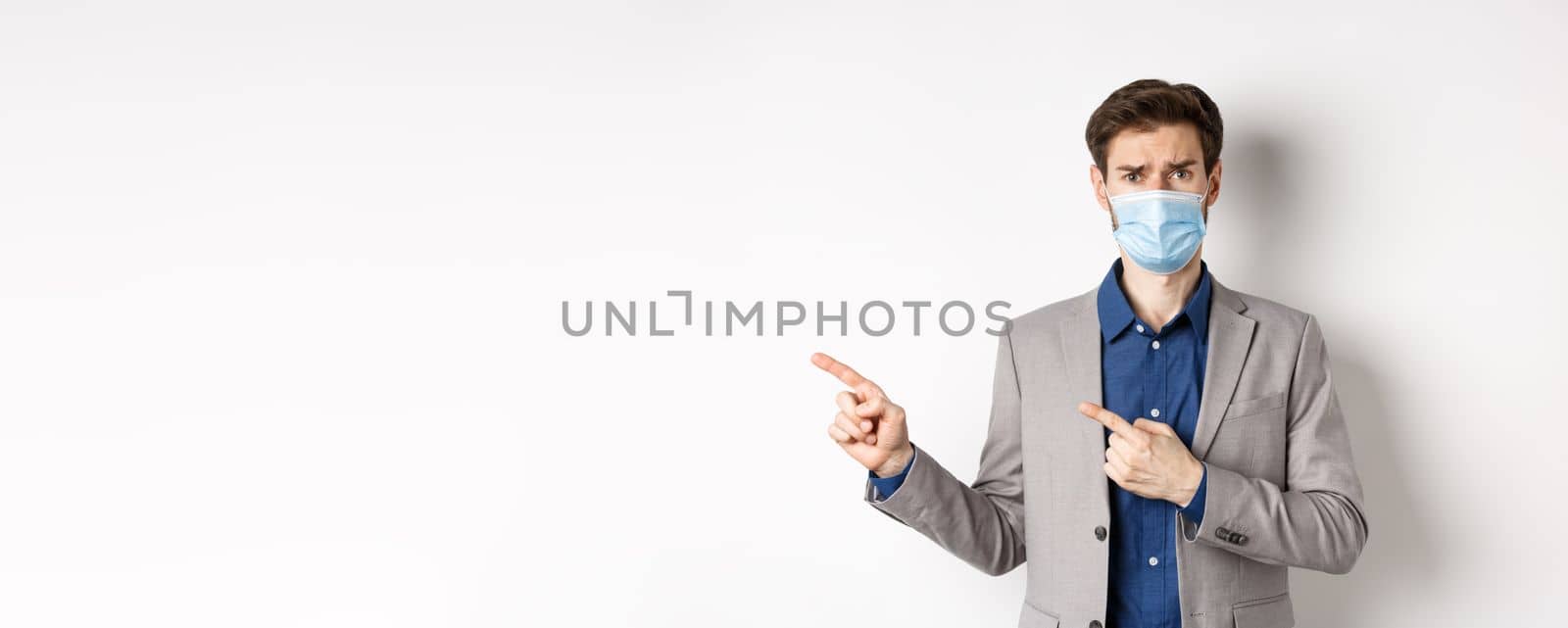 Covid-19, pandemic and business concept. Upset man in medical mask and office suit, frowning sad and pointing left at banner, standing on white background.