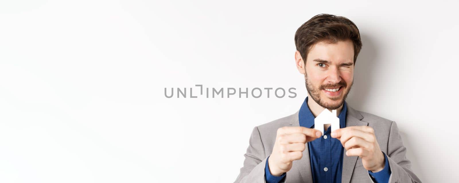 Real estate and insurance concept. Handsome man in suit winking and smiling, showing small paper house cutout, standing on white background by Benzoix