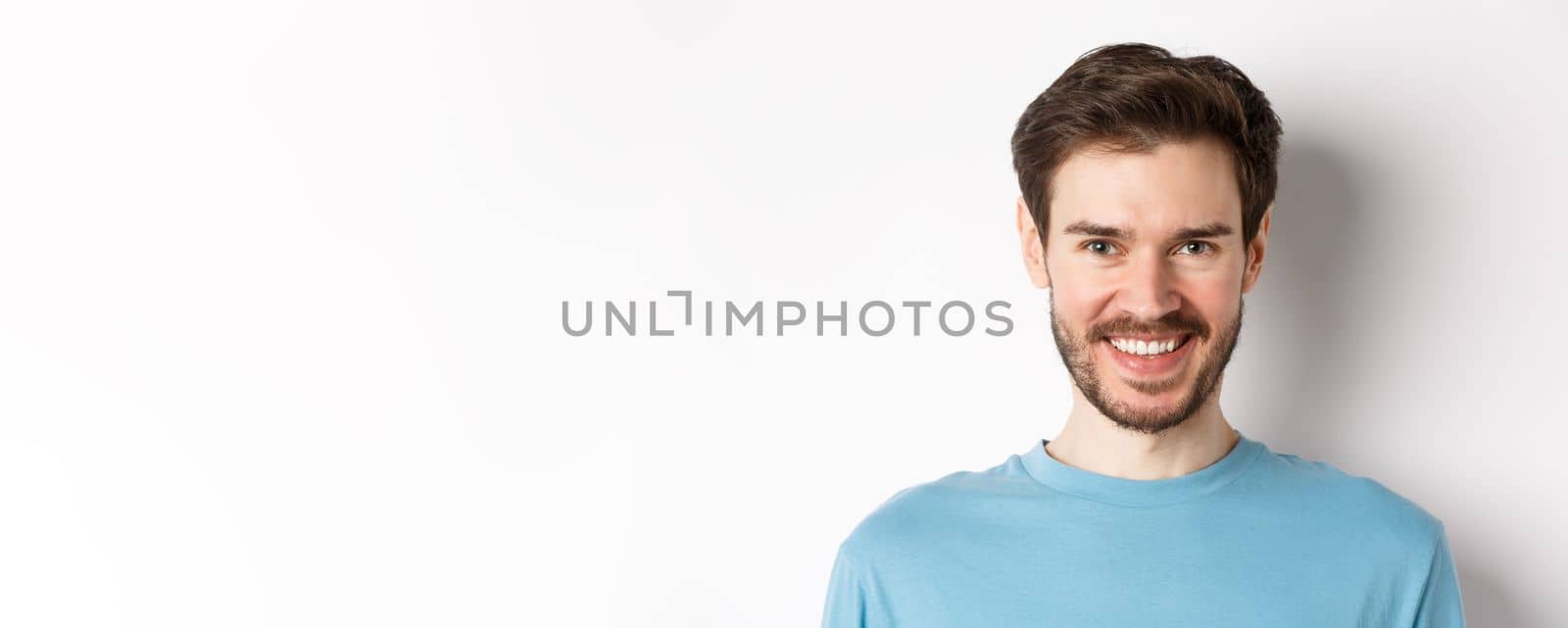 Close-up of handsome caucasian man smiling with white teeth, looking confident at camera, standing in blue shirt on white background by Benzoix