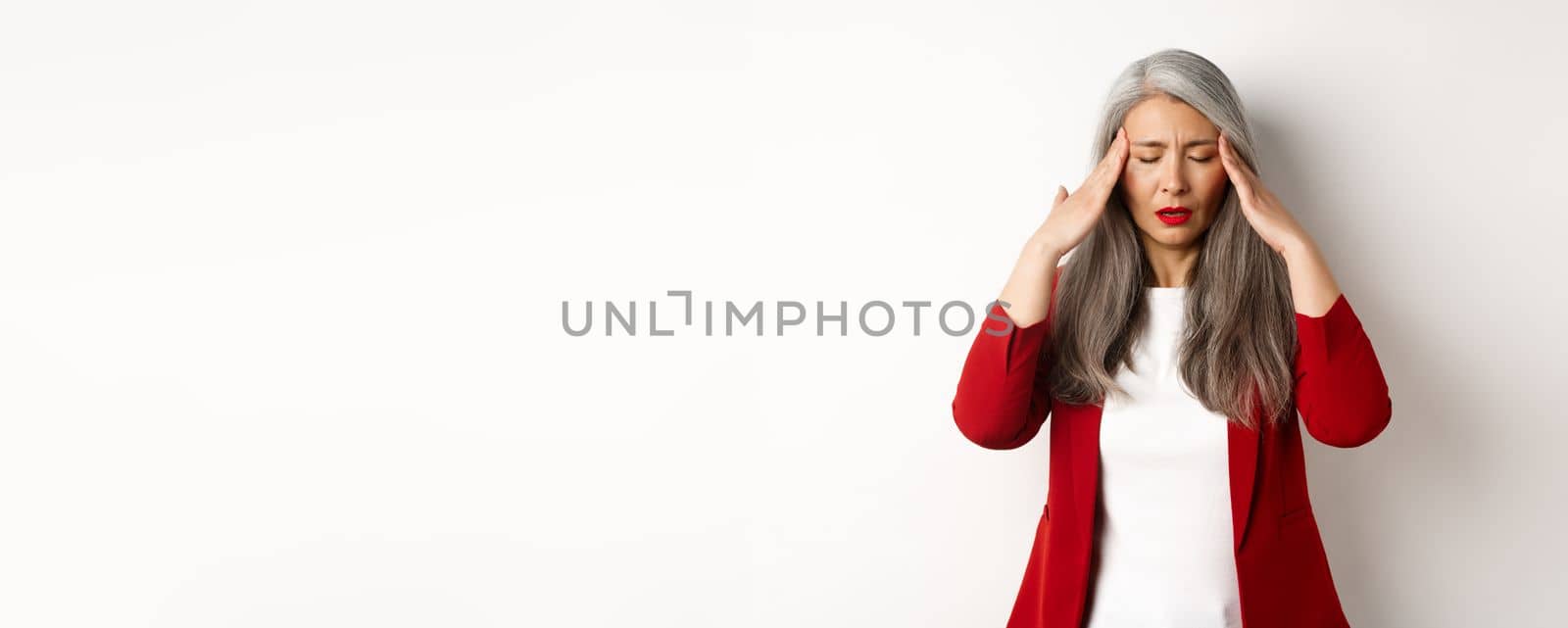 Tired senior businesswoman in red blazer having headache, touching head and feel sick, standing with migraine against white background.