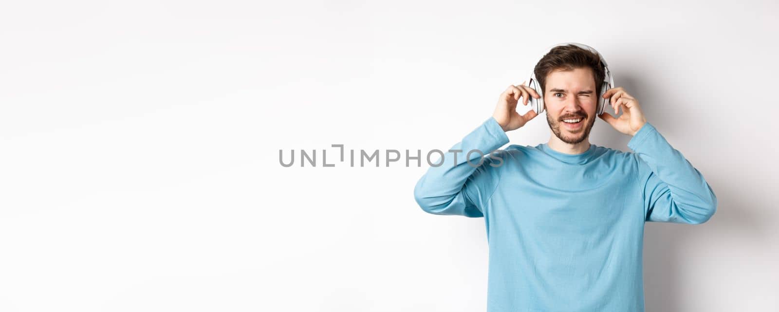 Cheerful guy wink at camera and smiling, listening music in wireless headphones, white background.