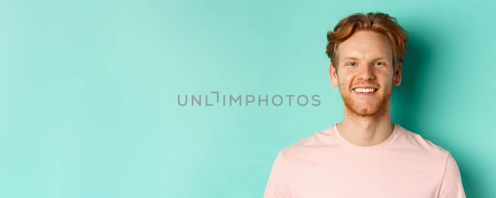 Close up of redhead bearded guy in pink t-shirt, smiling with white perfect teeth and looking at camera, standing over turquoise background by Benzoix