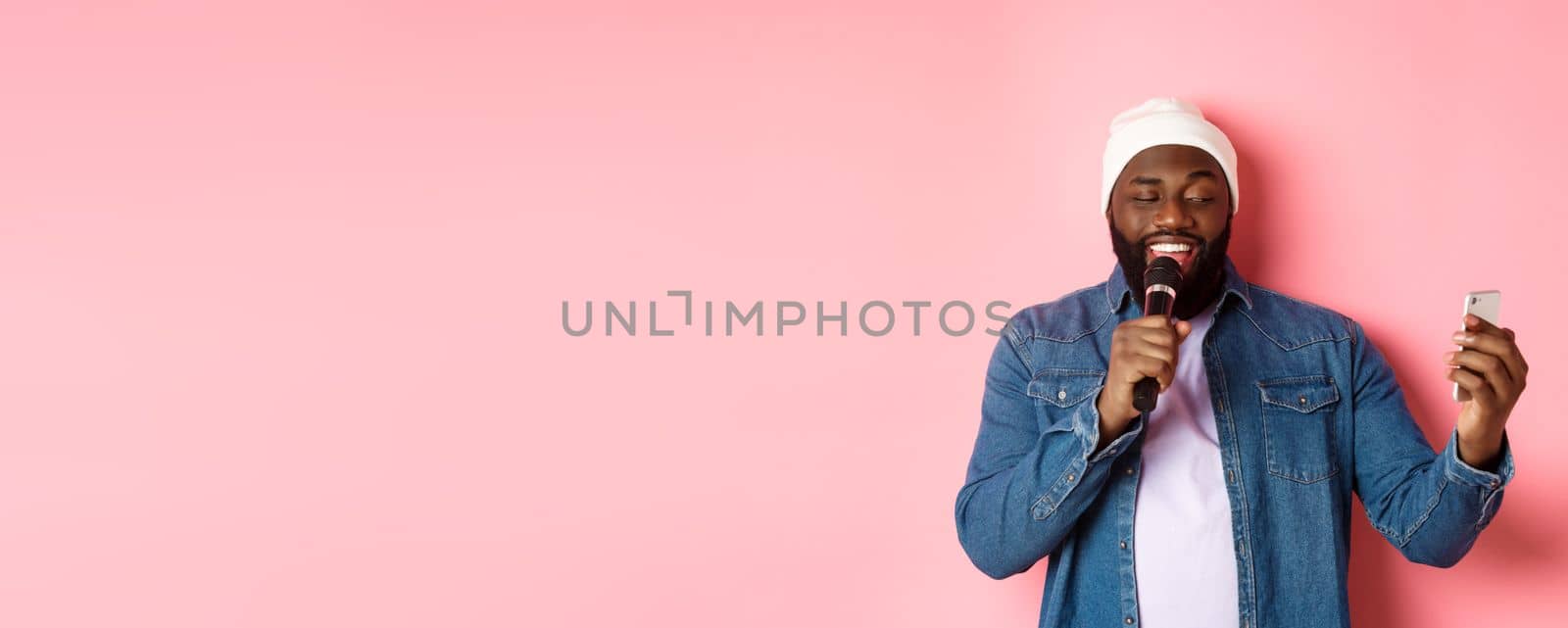 Handsome african-american man singing karaoke, reading lyrics on smartphone app and holding microphone, standing over pink background by Benzoix