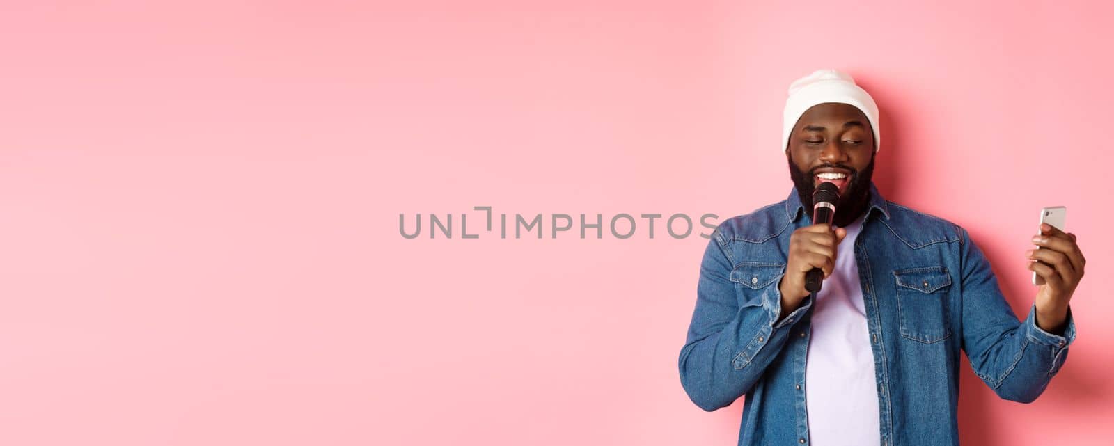 Handsome african-american man singing karaoke, reading lyrics on smartphone app and holding microphone, standing over pink background.