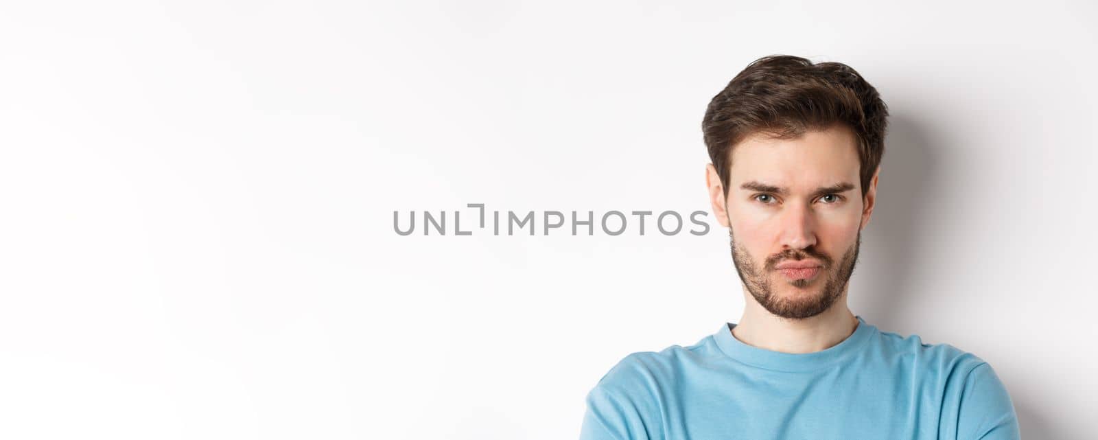 Close-up of grumpy young man grimacing, sulking and making offended face, standing upset over white background.